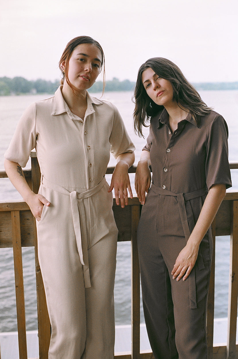 Two models standing up, leaning against a guardrail, looking at the camera, brown coloured jumpsuit, another is eggnog colour, brazilian crepe patou, river, lake in the background