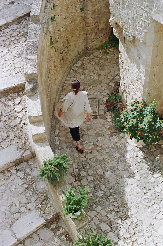 Model walking away in the distance into an alley. Plants, mediterrenean. Wearing sandals, black pants and a linen beije robe