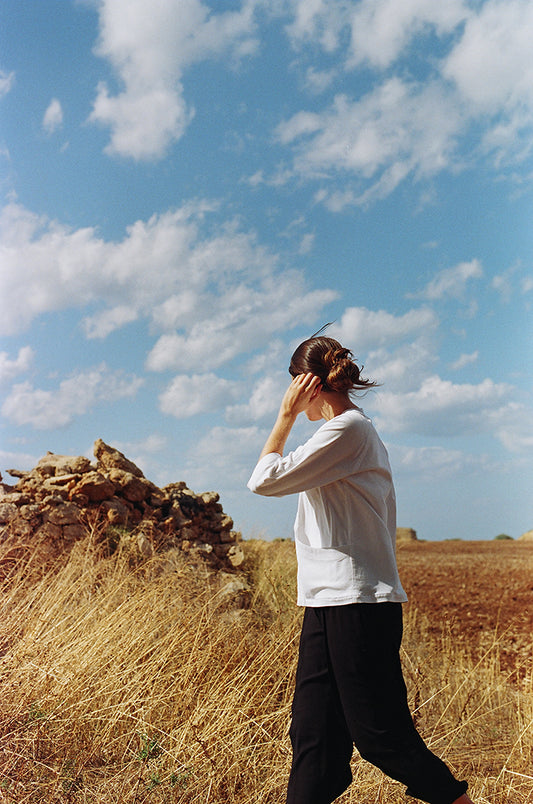 Model walking a field, blue sky, looking away. black pants, white linen Writer Jacket