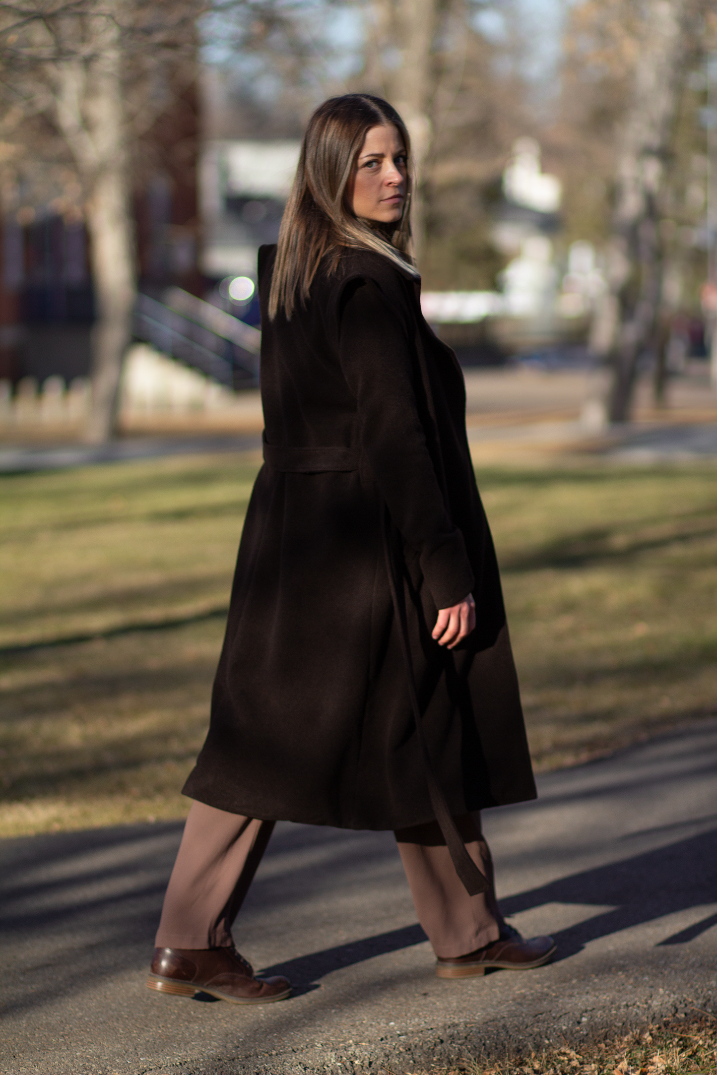 Model wearing The Designer Coat, a brown wool from southern Brazil on top of the Florist Jumpsuit, hot chocolate colour, with brown boots. Grass and trees in the background, model is walking away to the rightside of the picture, but with her face looking at the camera