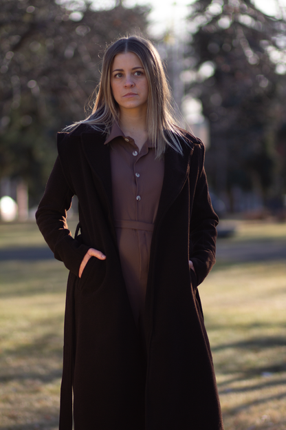 Model wearing The Designer Coat, a brown wool from southern Brazil on top of the Florist Jumpsuit, hot chocolate colour. Grass and trees in the background, model is facing the camera, looking up