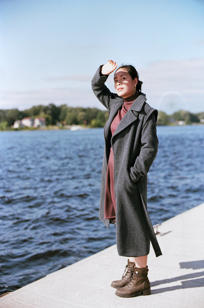 Model stanting up on a river dock, protecting her face against the sun, wearing boots and a cherry brown polyester dress, neoprene, with a gray wool jacket, covering down to her ankles.