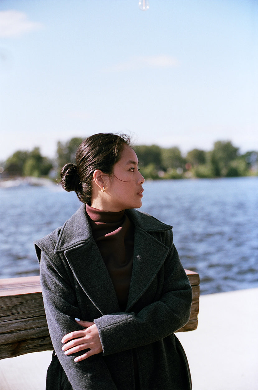 Model stanting up on a river dock, leaning on a wooden high table, wearing a cherry brown polyester dress, neoprene, with a gray wool jacket, covering down to her ankles.