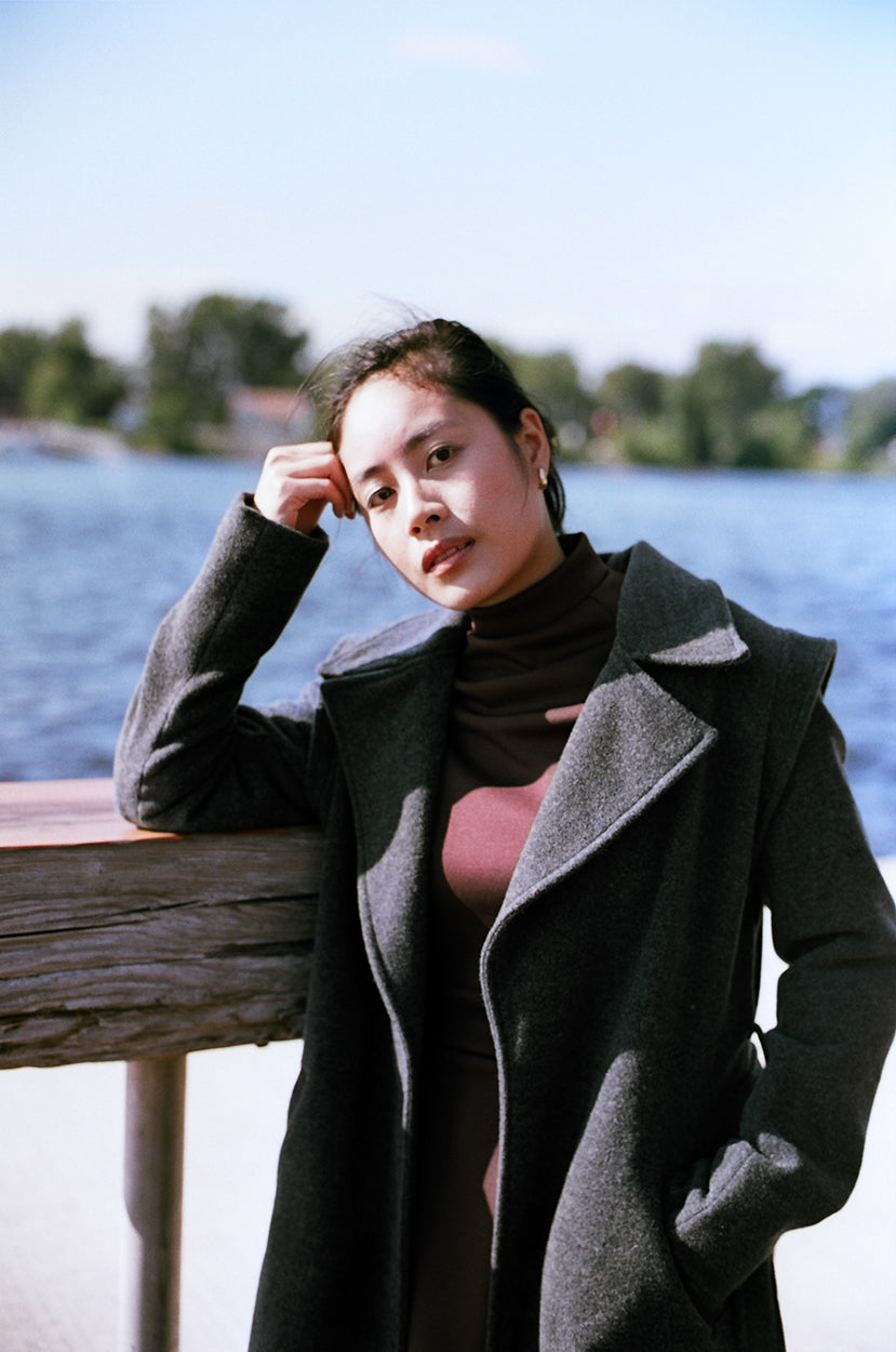 Model stanting up on a river dock, leaning on a wooden high table, looking at the camera, wearing a cherry brown polyester dress, neoprene, with a gray wool jacket, covering down to her ankles.