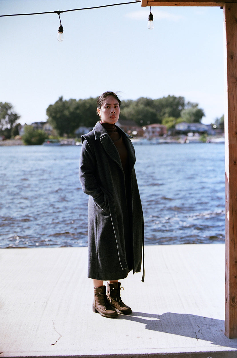 Model stanting up on a river dock, llooking at the camera, wearing boots and a cherry brown polyester dress, neoprene, with a gray wool jacket, covering down to her ankles.