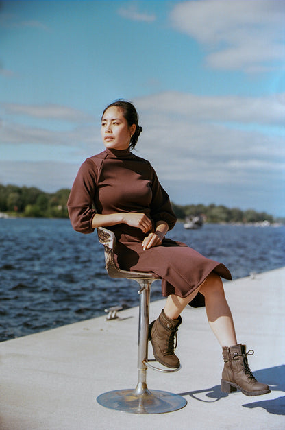 Model sitting on s silver chair, on a river dock, looking at the river, wearing boots and a cherry brown polyester dress, neoprene