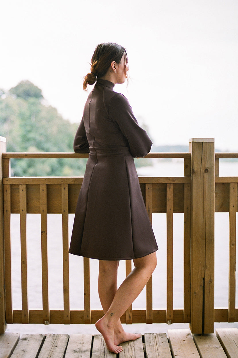 Model leaning on the guardrail, looking at a river, trees in the background, wearing a polyester dress, neoprene, barefeet.