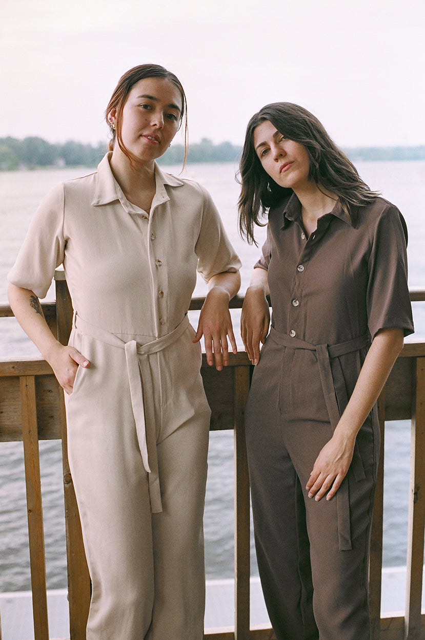Two models standing up, leaning against a guardrail, looking at the camera, brown coloured jumpsuit, another is eggnog colour,  brazilian crepe patou, river, lake in the background