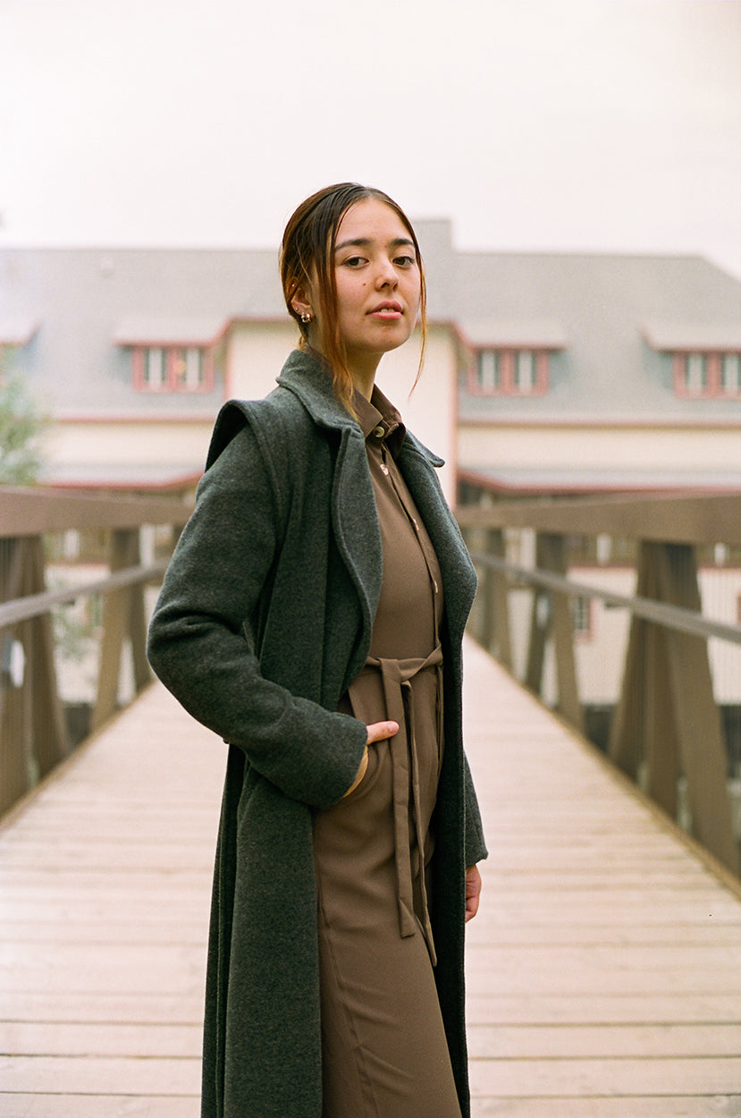 Model looking at the camera, standing up on a overpass, boathouse in the background, wearing brown crepe patou jumpsuit, with a gray wool jacket, covering down to her ankles.