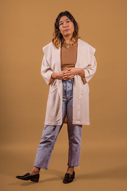 Model stanting up, looking at the camera on a beije background. Standing up, brown shoes, blue jeans, light brown blouse, wearing a beije linen robe