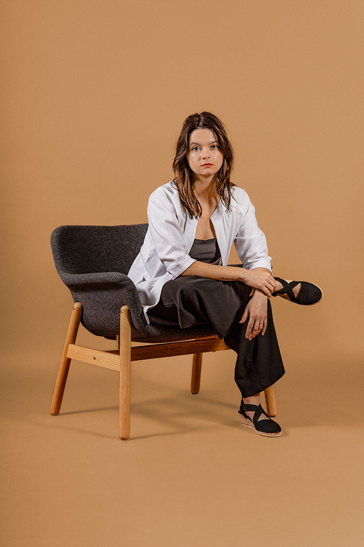 Brunete model on a beije background sitting on a grey chair, looking at the camera. black pants, white linen "writer Jacket"