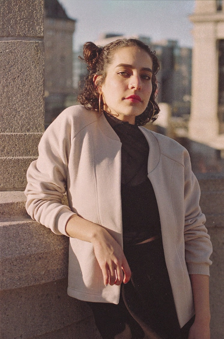 Model on leaning on a wall on a sunny day, black pants, black blouse, beije cottom fleece jacket